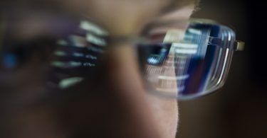 Portait of young hacker with computer screen reflecting in the man's glasses.